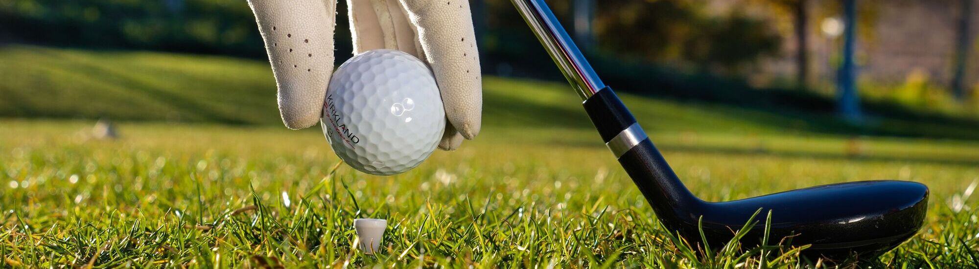A close-up of a gloved hand placing a golf ball on a tee, with a golf club positioned nearby, set on a grassy course.
