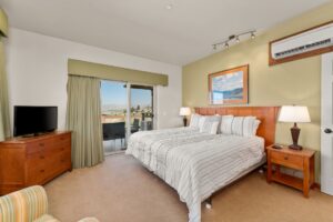 A bright bedroom with a large bed, wooden furniture, two bedside lamps, a TV on a dresser, and sliding glass doors leading to a balcony with a scenic mountain view.