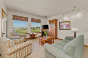 A cozy living room with large windows showing a scenic view, two armchairs, a green sofa, a wooden coffee table with flowers, a TV on a cabinet, and wall art and lamp for decor.