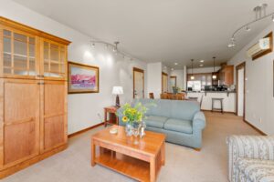 A bright living room with a wooden coffee table, light blue couch, armchair, and a bouquet of flowers. A wooden cabinet, framed artwork, and a kitchen with barstools are visible in the background.