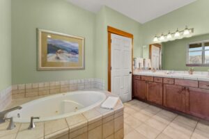 Spacious bathroom with a large corner bathtub, light green walls, wooden cabinets, double sink vanity, wall mirror, towel rack, and framed landscape artwork above the tub.