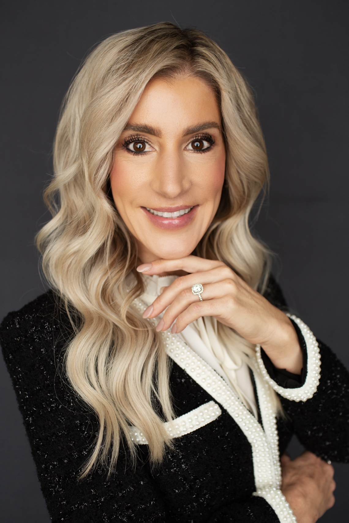 A woman with long, wavy blonde hair smiles at the camera. She is wearing a black jacket with white trim and pearls, a white blouse, and a diamond ring on her finger, posed against a dark background.