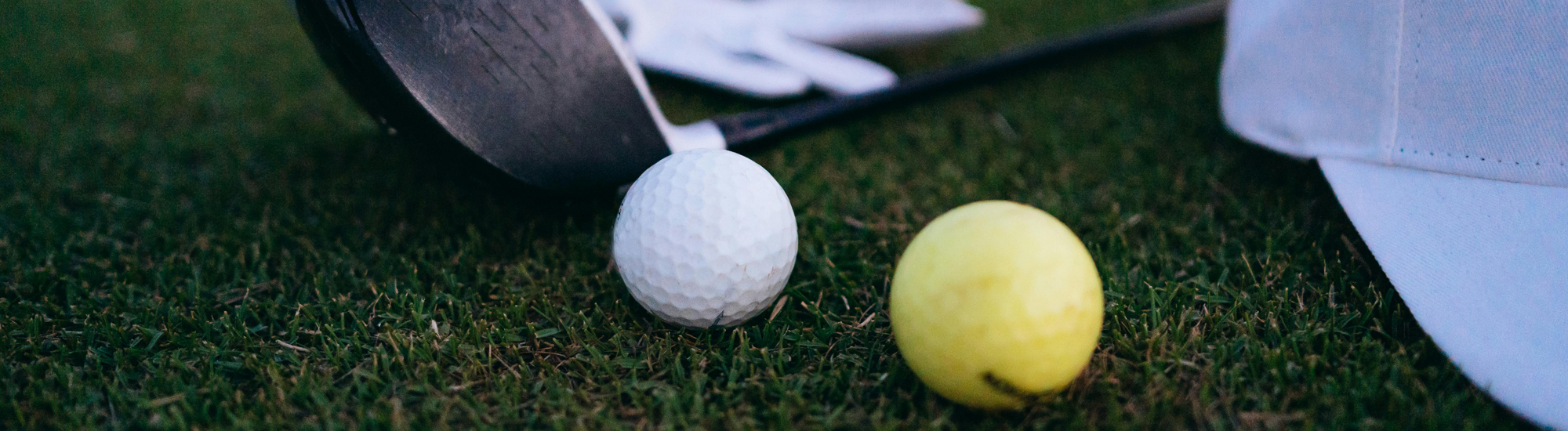 Close-up of a white golf ball and a yellow golf ball on grass, with a golf club head, white glove, and part of a white cap visible in the background.