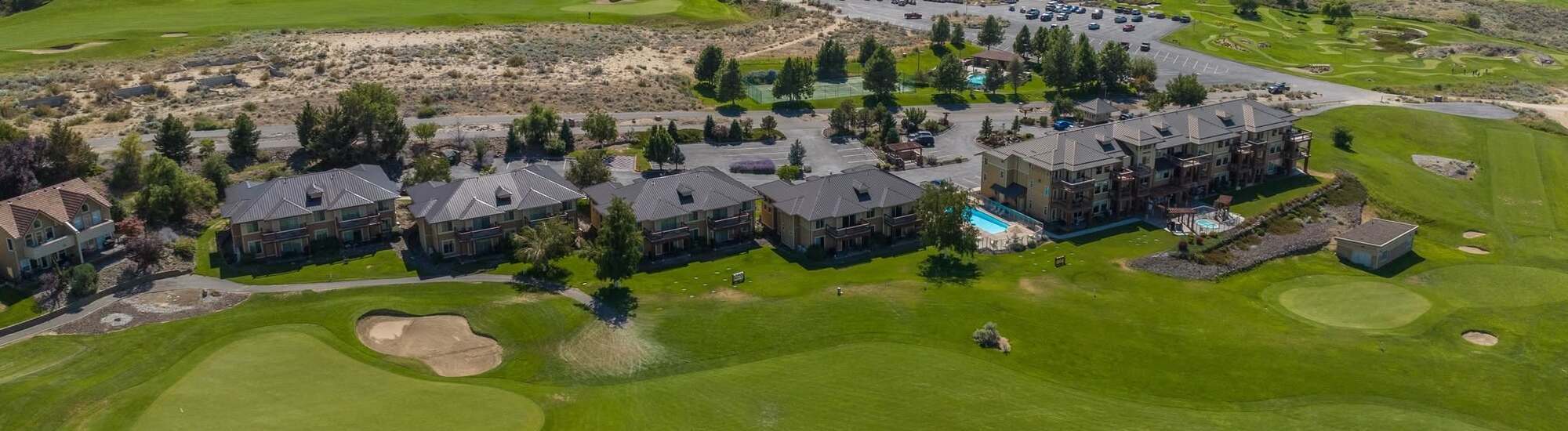 Aerial view of a row of residential buildings beside a golf course, with green fairways, sand bunkers, a swimming pool, and parked cars visible under a clear sky.