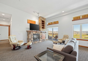 A bright living room with a stone fireplace, mounted TV, gray sofa, two cream armchairs, large windows, and a glass door opening to a scenic mountain view.
