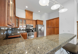 Modern kitchen with wooden cabinets, stainless steel appliances, a granite countertop bar with a single stool, built-in microwave and oven, and three pendant lights hanging above the counter.