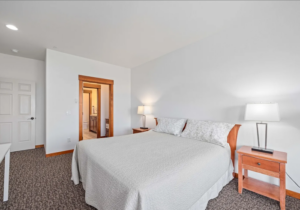 A bright bedroom with a large bed covered in a white quilt, two wooden nightstands with lamps, beige carpet, and a doorway leading to a bathroom. The walls are white and the space looks tidy and minimal.