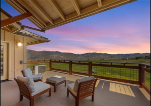 A covered balcony with cushioned wooden chairs and an ottoman overlooks a scenic landscape of rolling hills and grassy fields under a pink and purple sunset sky.