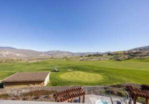 A wide view of a golf course with a green, rolling hills and mountains in the distance under a clear blue sky. In the foreground, there’s a fenced patio area and pergolas.