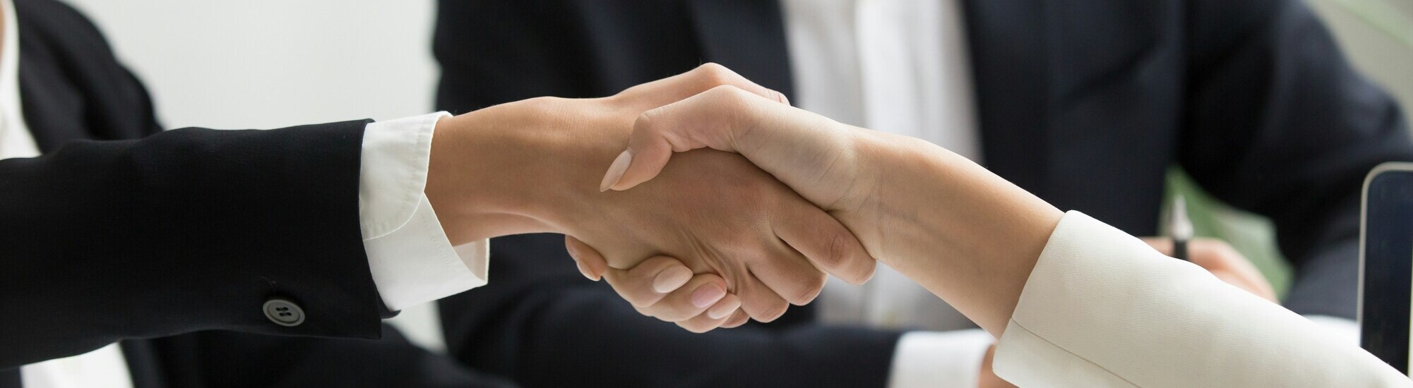 Two people in business attire shaking hands, with another person sitting in the background. The image suggests a professional or business meeting or agreement.