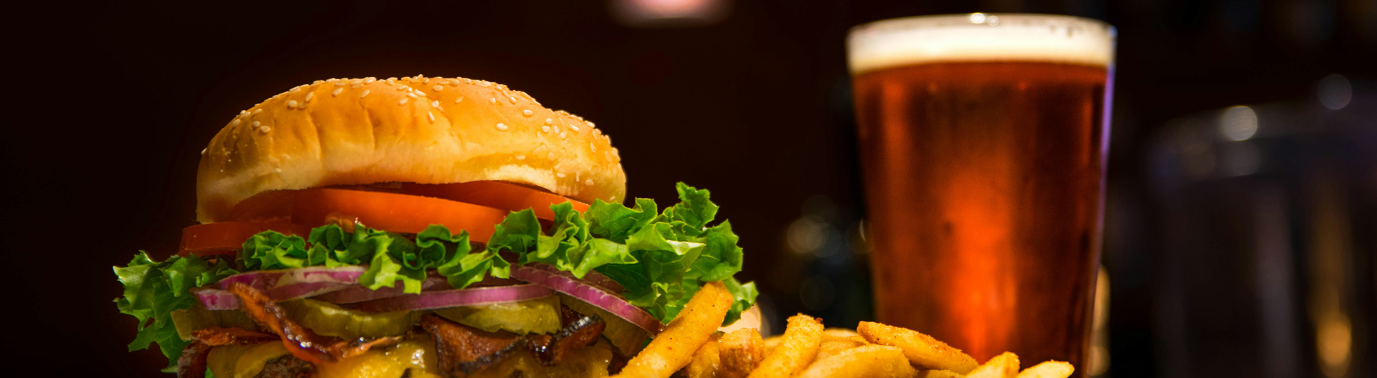 A close-up of a cheeseburger with lettuce, tomato, pickles, red onion, and bacon, served with a side of French fries. A glass of beer is in the background, all set against a dark, blurred backdrop.