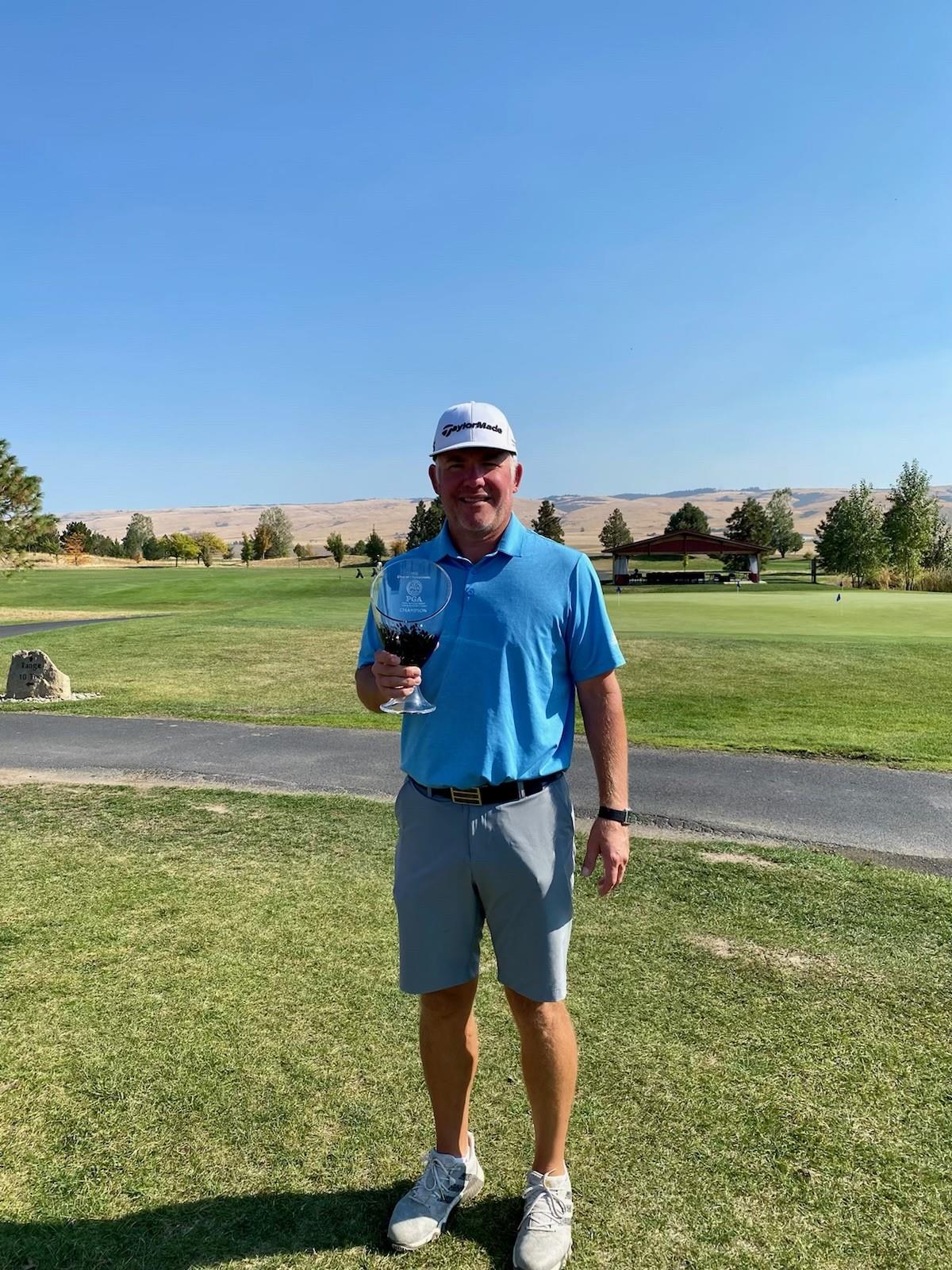A man wearing a blue polo shirt, gray shorts, white cap, and sunglasses stands on a golf course holding a trophy. The sky is clear and trees, grass, and hills are visible in the background.