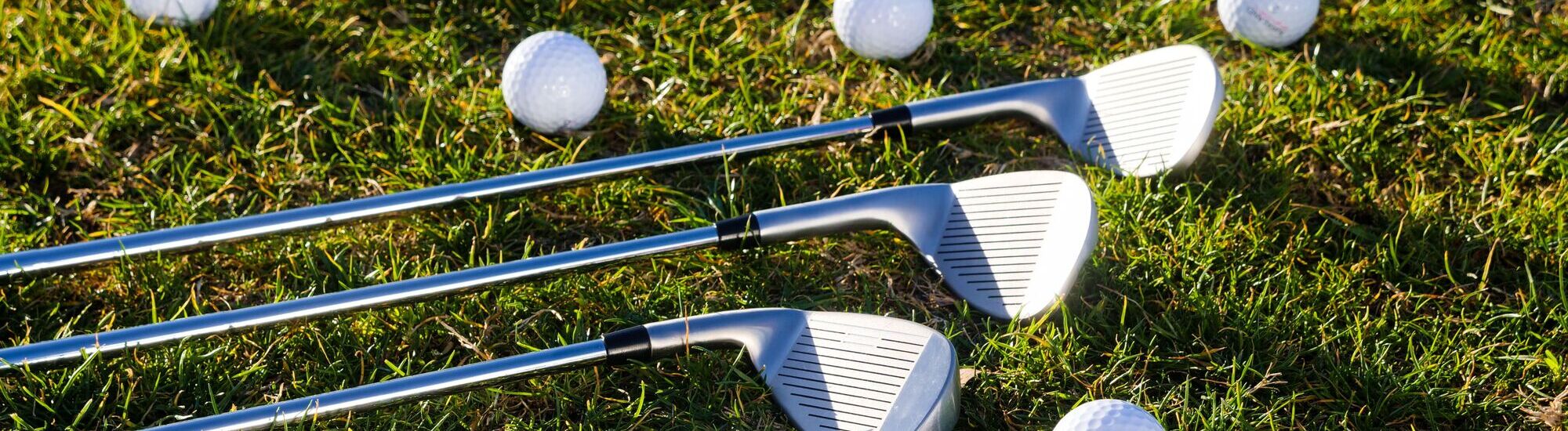 Three golf clubs lie on green grass with several white golf balls scattered around them.