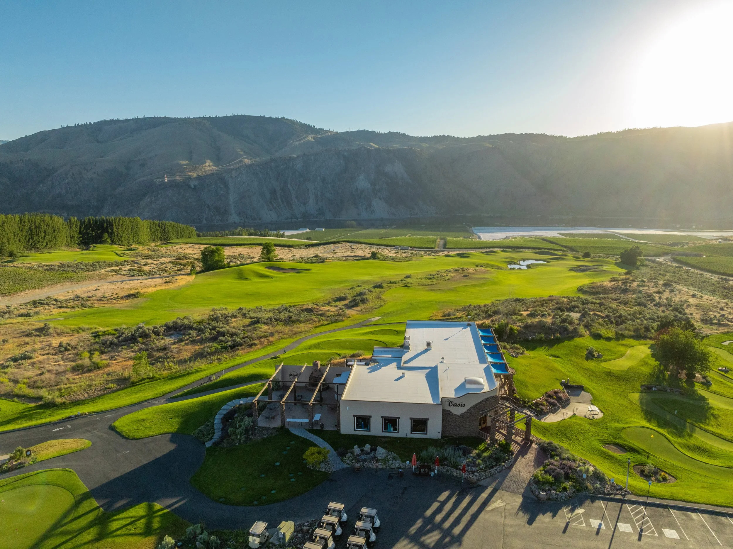 Aerial view of a golf course with a clubhouse in the foreground, surrounded by green fairways, rolling hills, and distant mountains under a clear blue sky at sunset.